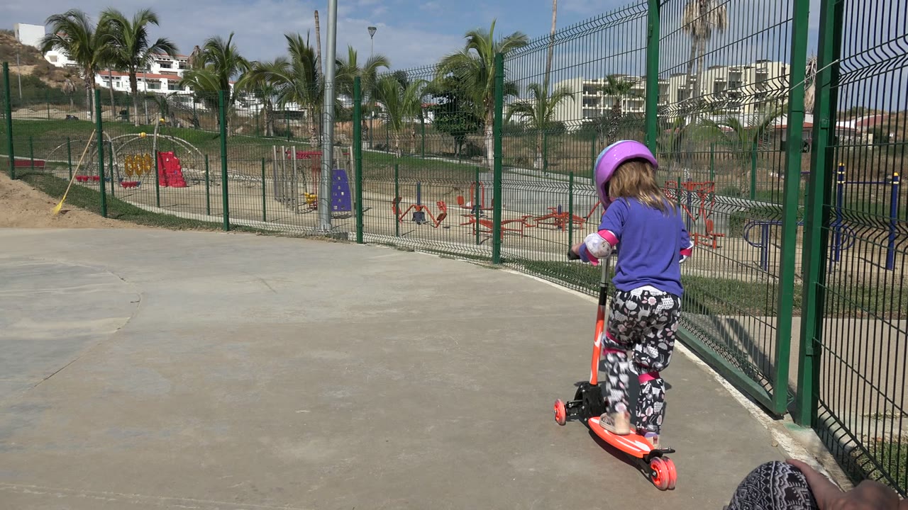 Helmeted child riding an orange scooter beside a green playground fence.