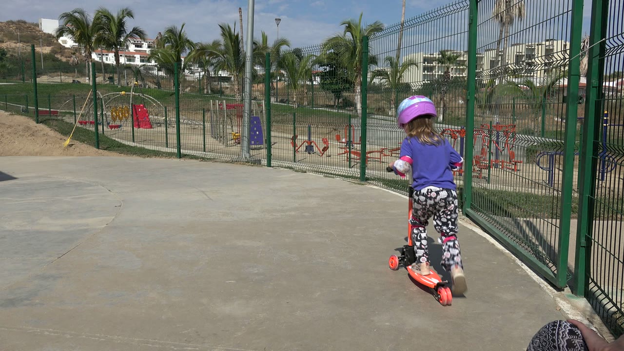 The child rides the orange scooter farther from the camera in Los Cabos, passing the green fence with red and purple playground structures and palm trees in the background.