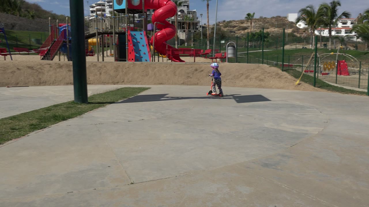 A helmeted child rides a small scooter on a concrete path at a Los Cabos playground, with red slides and sandy play areas behind them.