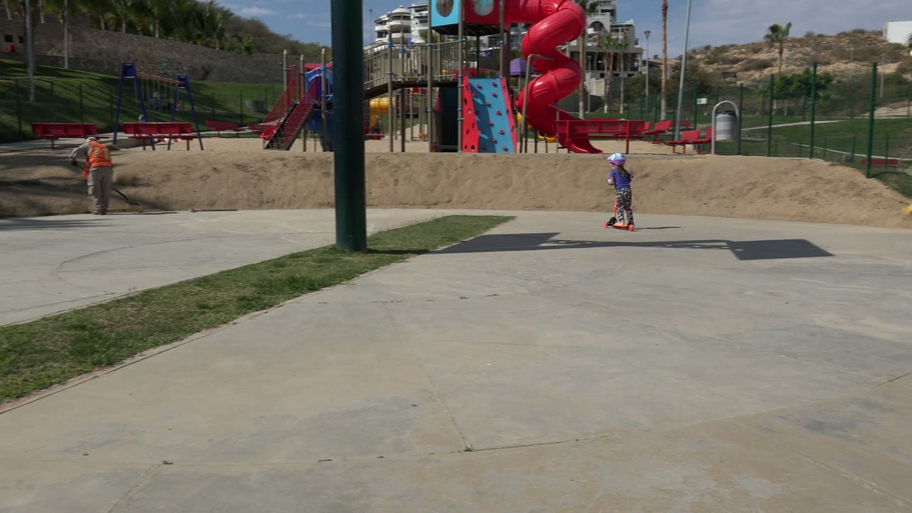 A worker in an orange vest shovels dirt beside the concrete walkway at a Los Cabos playground while red benches and play structures sit in the background.