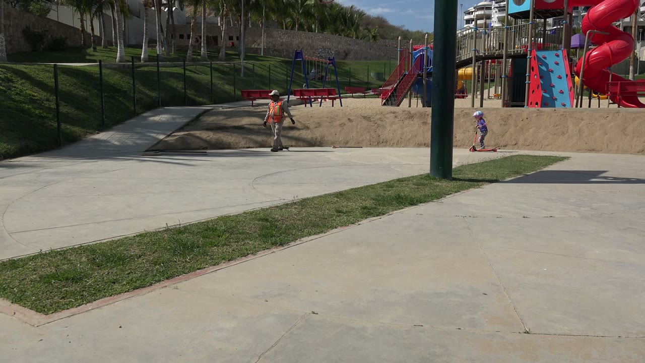 A child rides a red scooter on a concrete path in a Los Cabos park, with sandy playground ground, red play equipment, grass, palms, and buildings behind.