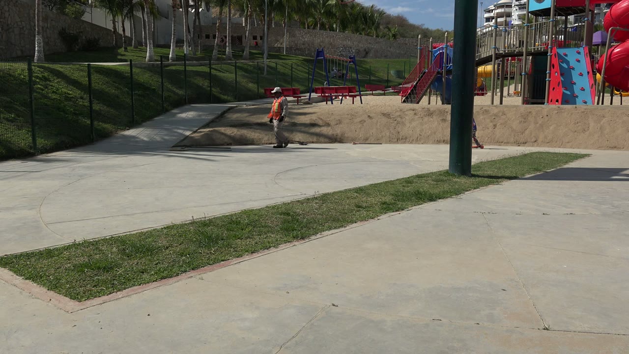 The child continues across the concrete walkway in the Los Cabos park, passing near a green pole with red slides and climbing panels in the background.