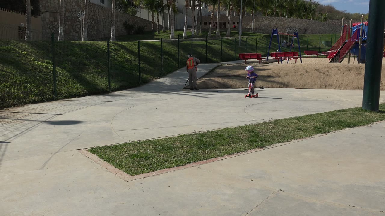 A child rides a red scooter across a concrete path in a Los Cabos park while an adult walks farther back near grass and playground equipment.