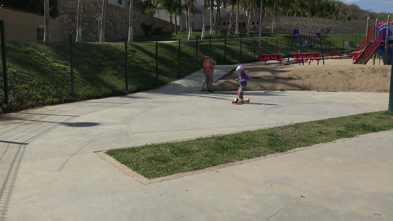 The child continues riding the red scooter through the Los Cabos park, with an adult walking behind and red benches and a playground slide across the sandy area.