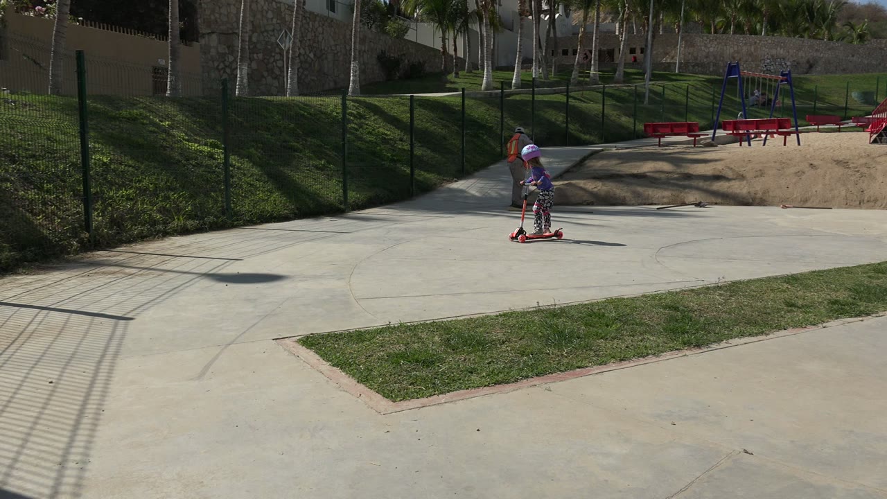 Child on scooter moving right across an open park path.