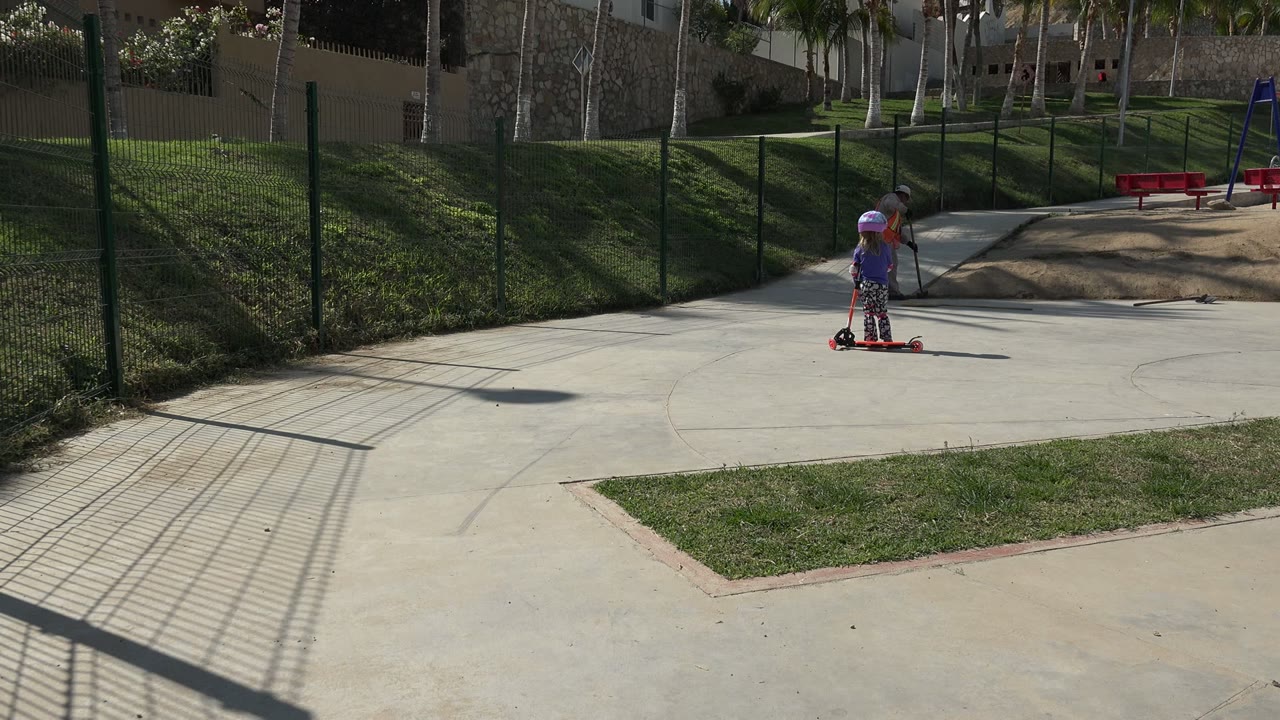 A helmeted child stands on a red scooter on a concrete park path in Los Cabos, Mexico, with a fenced lawn, benches, swings, and an adult farther back near the play area.