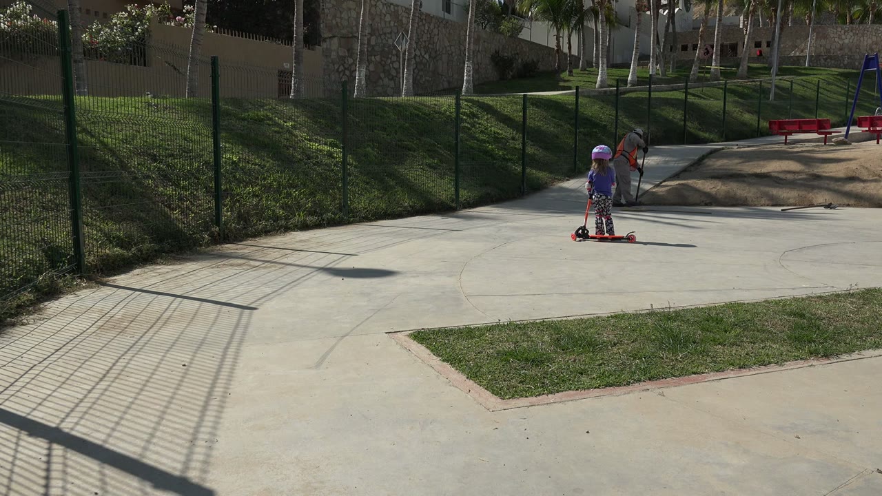 The child remains on the red scooter in the sunlit Los Cabos park while an adult in an orange vest bends near the sandy play area behind them.