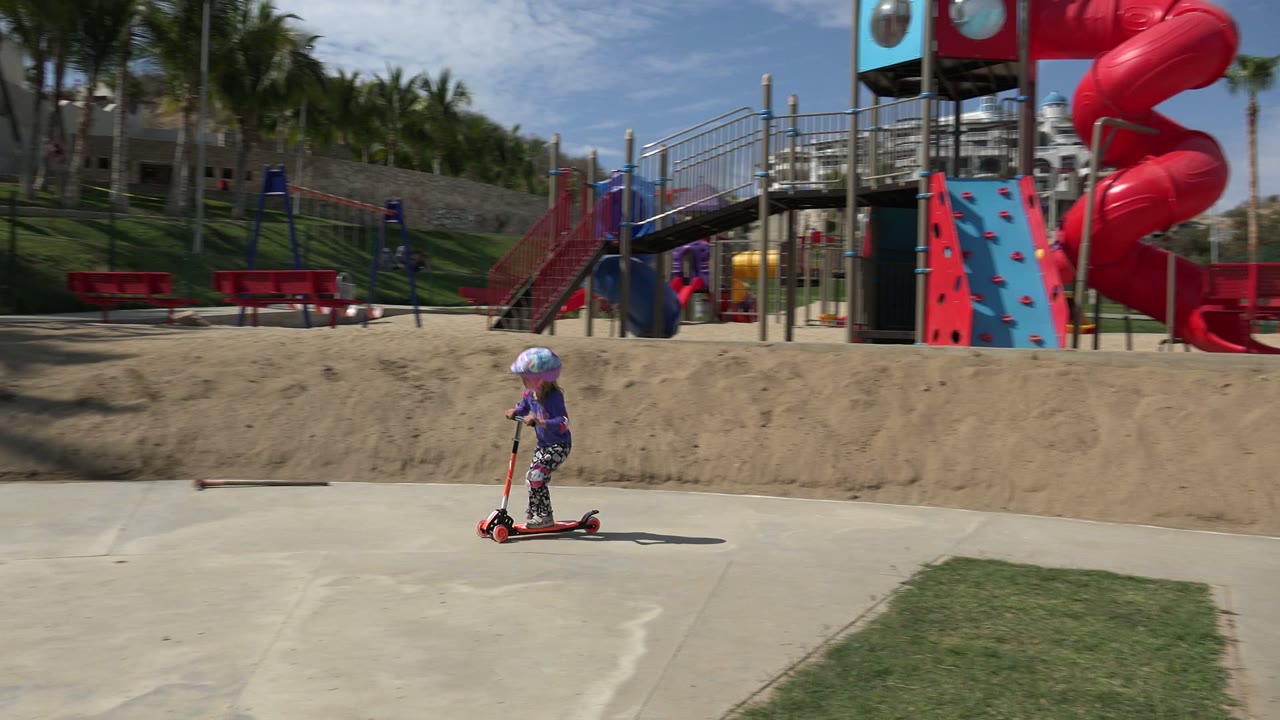 Young girl in a helmet rides a red scooter across a playground walkway.