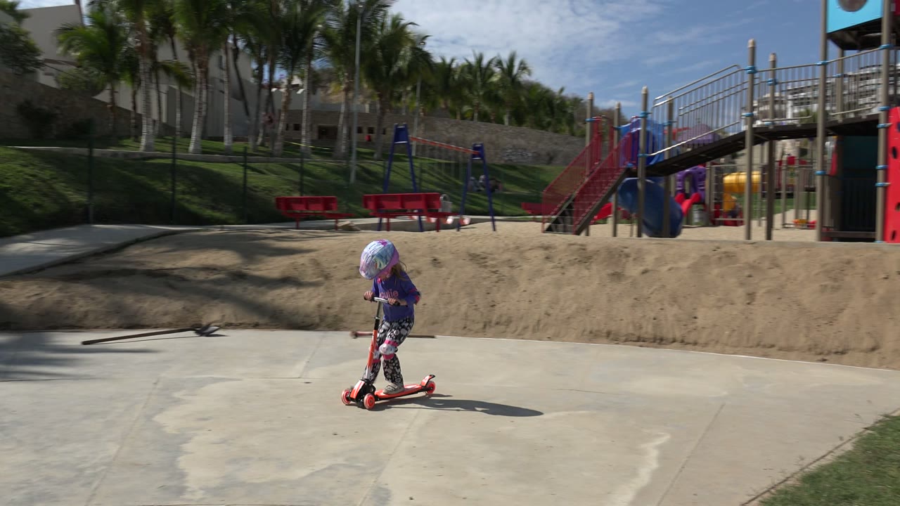 The helmeted child continues across the concrete in Los Cabos, Mexico, with a red slide, climbing structure, red benches, sand, and palm trees in the background.