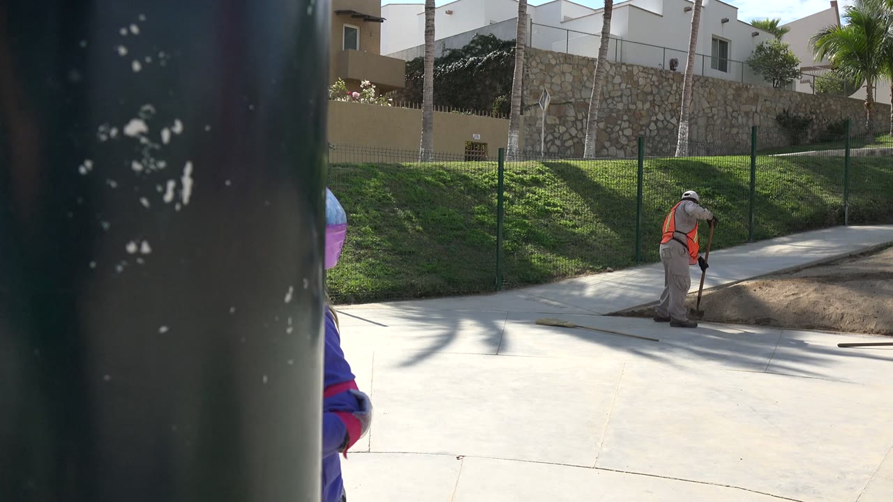 Child partly hidden by a dark post while a maintenance worker stands near a paved path.