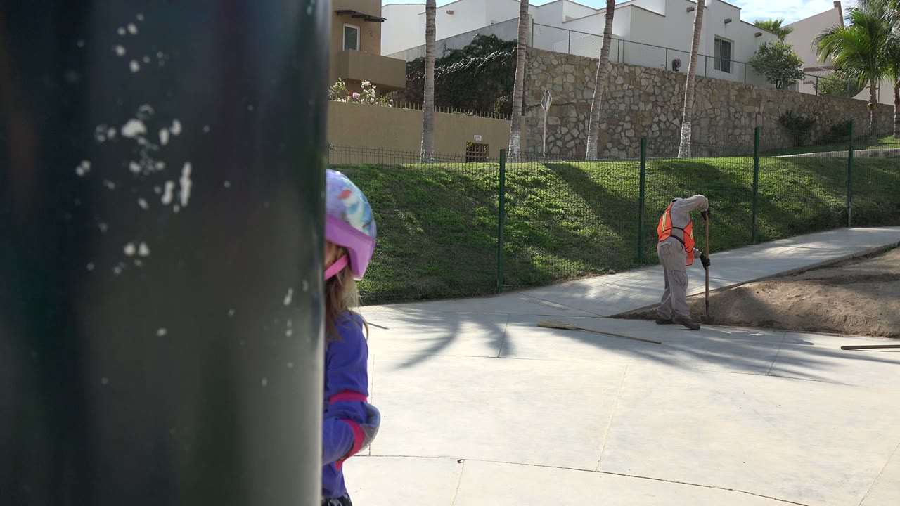 In Los Cabos, Mexico, the worker bends over the dirt beside the walkway while the helmeted child remains partially hidden at the left edge behind a dark post.