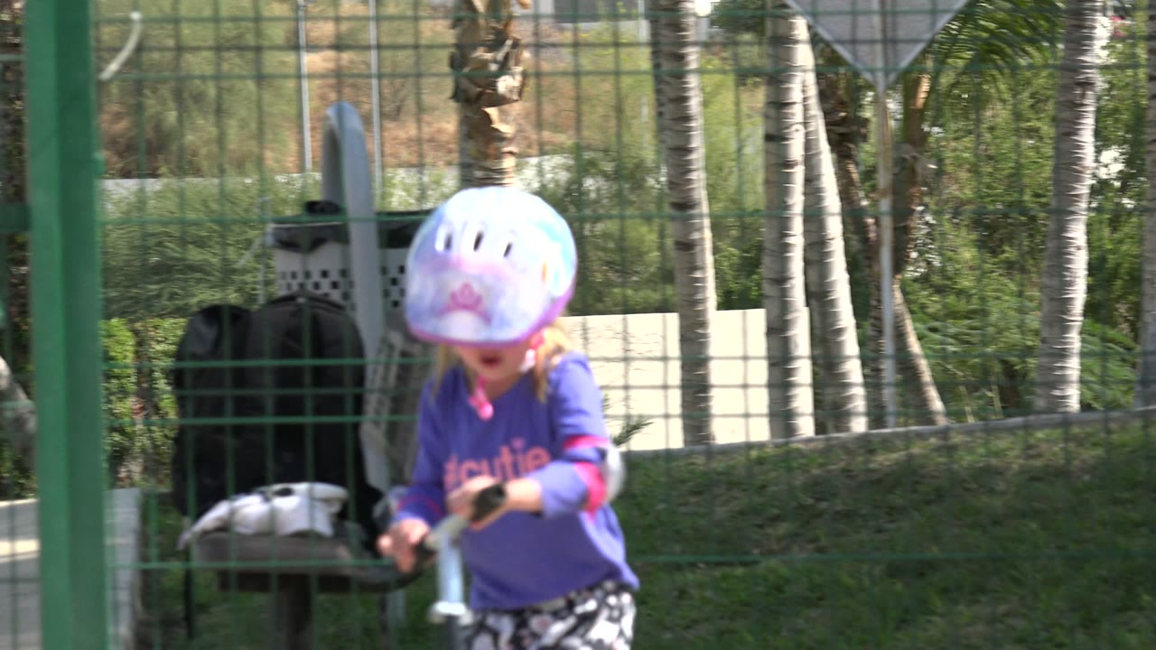 Blonde child in helmet rides a scooter behind a green fence.