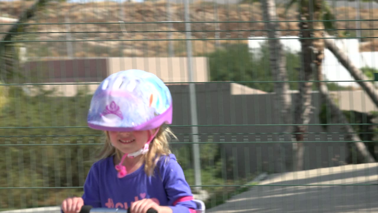 Young blonde girl in a helmet riding a scooter near a playground fence.