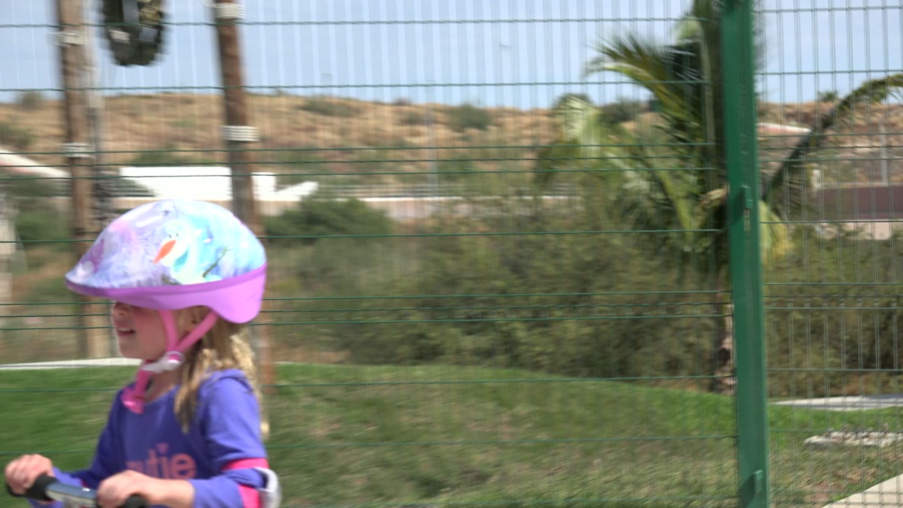 The girl moves farther across the Los Cabos playground, still close enough for her helmet, face, shirt, and scooter handlebar to be visible.