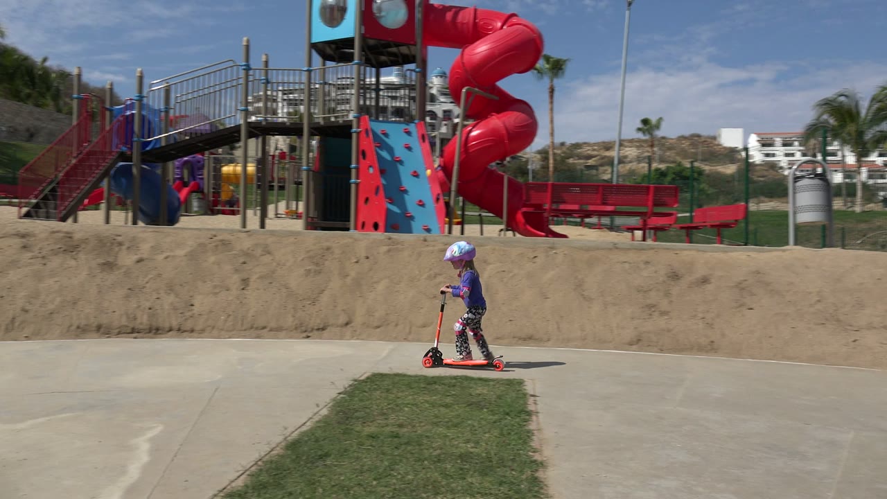 A helmeted child rides a red scooter across a paved park path in Los Cabos, Mexico, with a red slide and climbing wall behind them.
