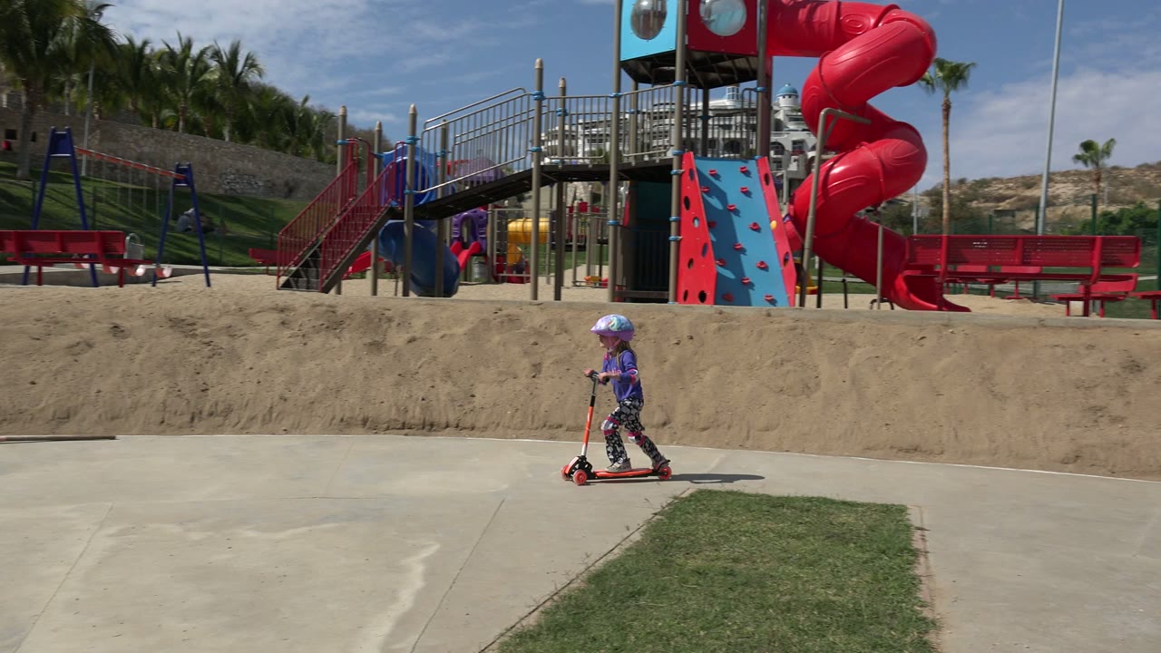 The child moves farther right on the scooter in the Los Cabos park, with red benches, palm trees, sandy ground, and playground stairs in the background.