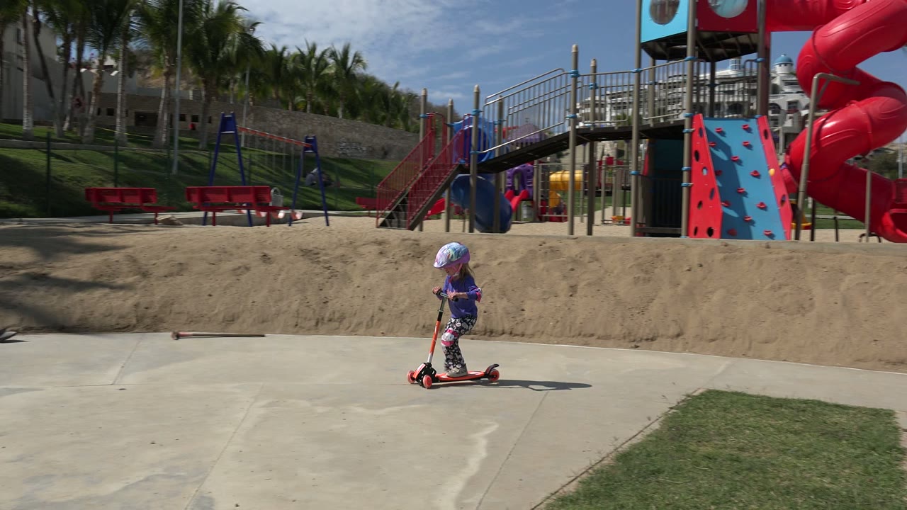 Child rides scooter past red playground equipment and palm trees.