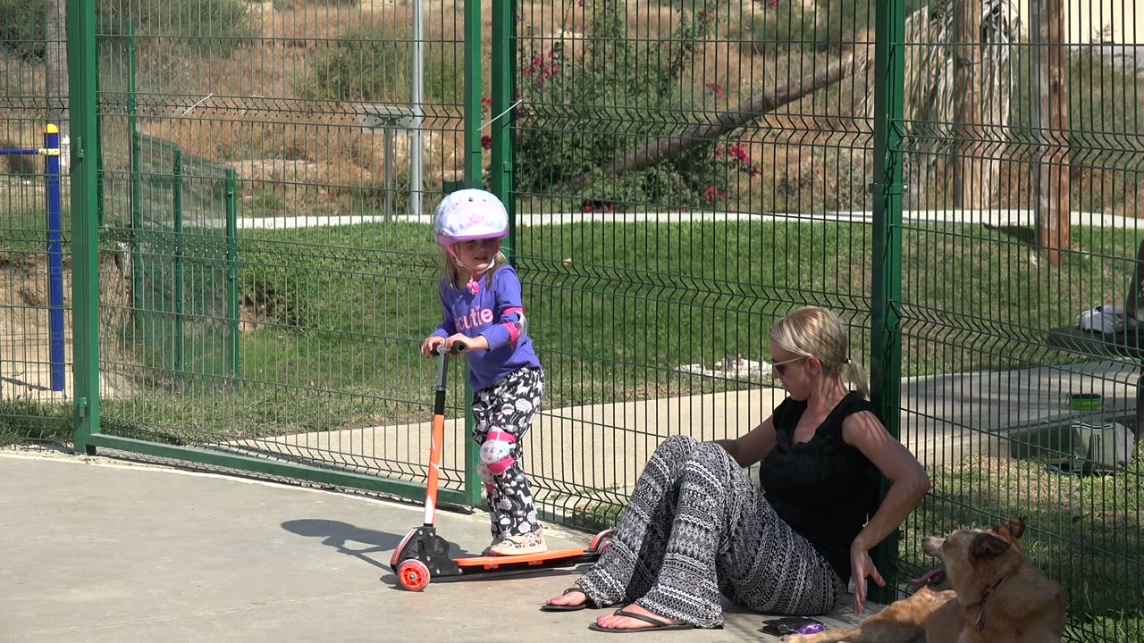 Young girl on a scooter beside a seated woman and dog.