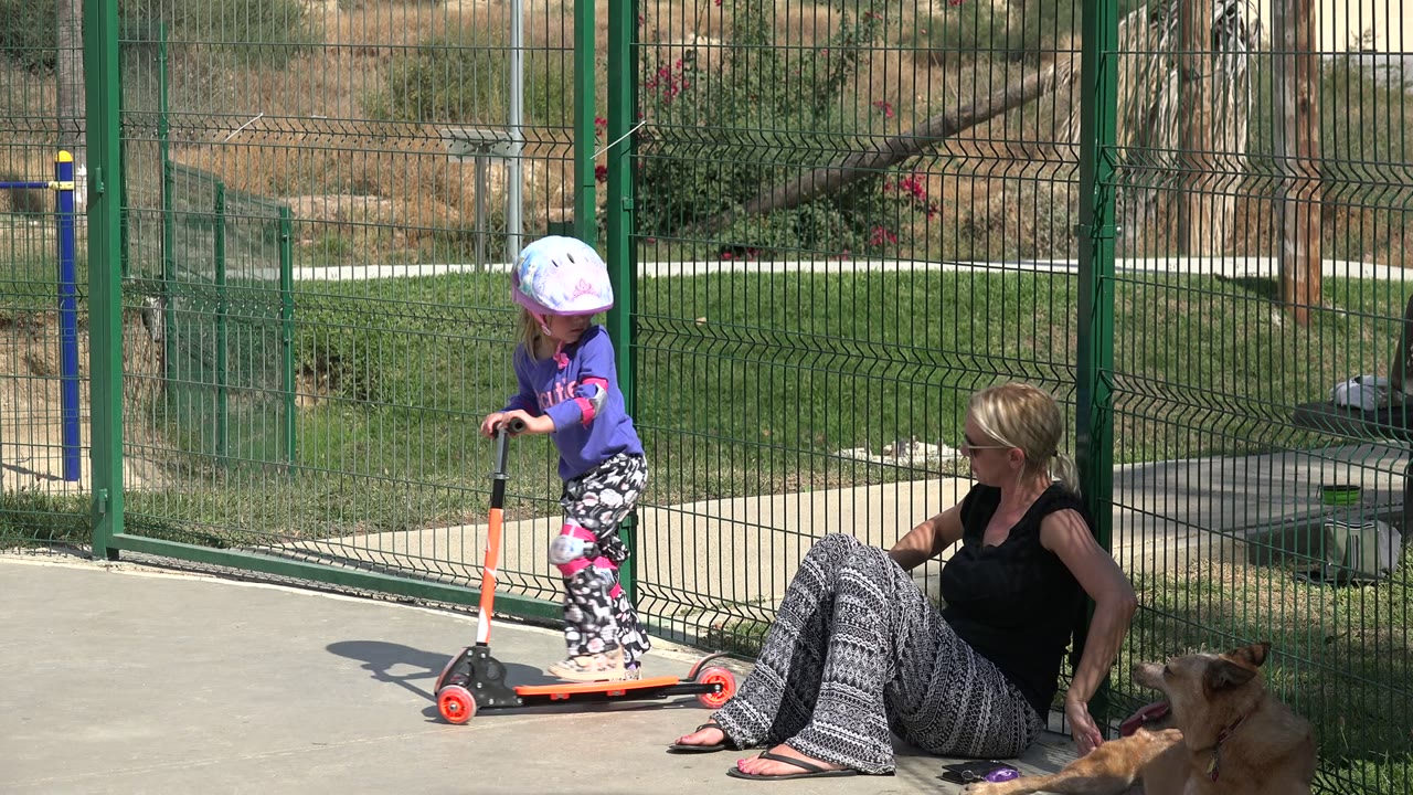 In Los Cabos, Mexico, the young girl pushes forward on the orange scooter as her mother sits to the right near the fence and the dog rests at the edge of frame.