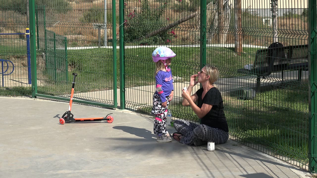 In Los Cabos, the mother kneels and lifts a small white item toward the helmeted child, with the scooter parked behind them on the concrete.