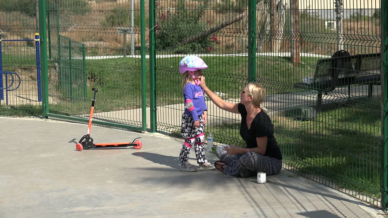 In Los Cabos, the young girl remains beside the scooter as the seated woman looks up at her, with a bench and grass behind the fence.