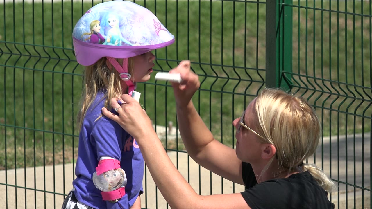 Woman applies sunscreen to a helmeted child beside a green fence.