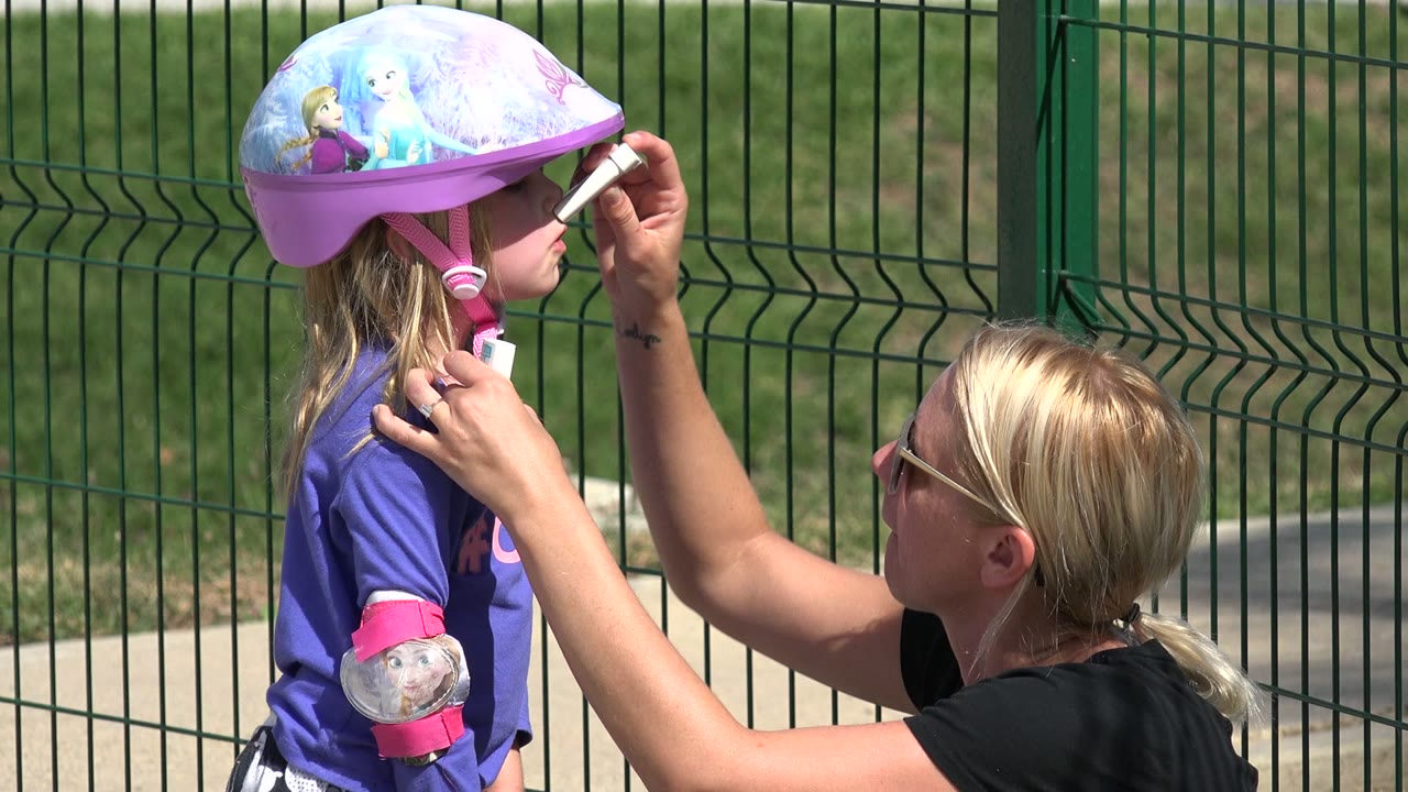 In Los Cabos, Mexico, the woman raises the sunscreen applicator close to the child's face while steadying the pink helmet strap with her other hand.