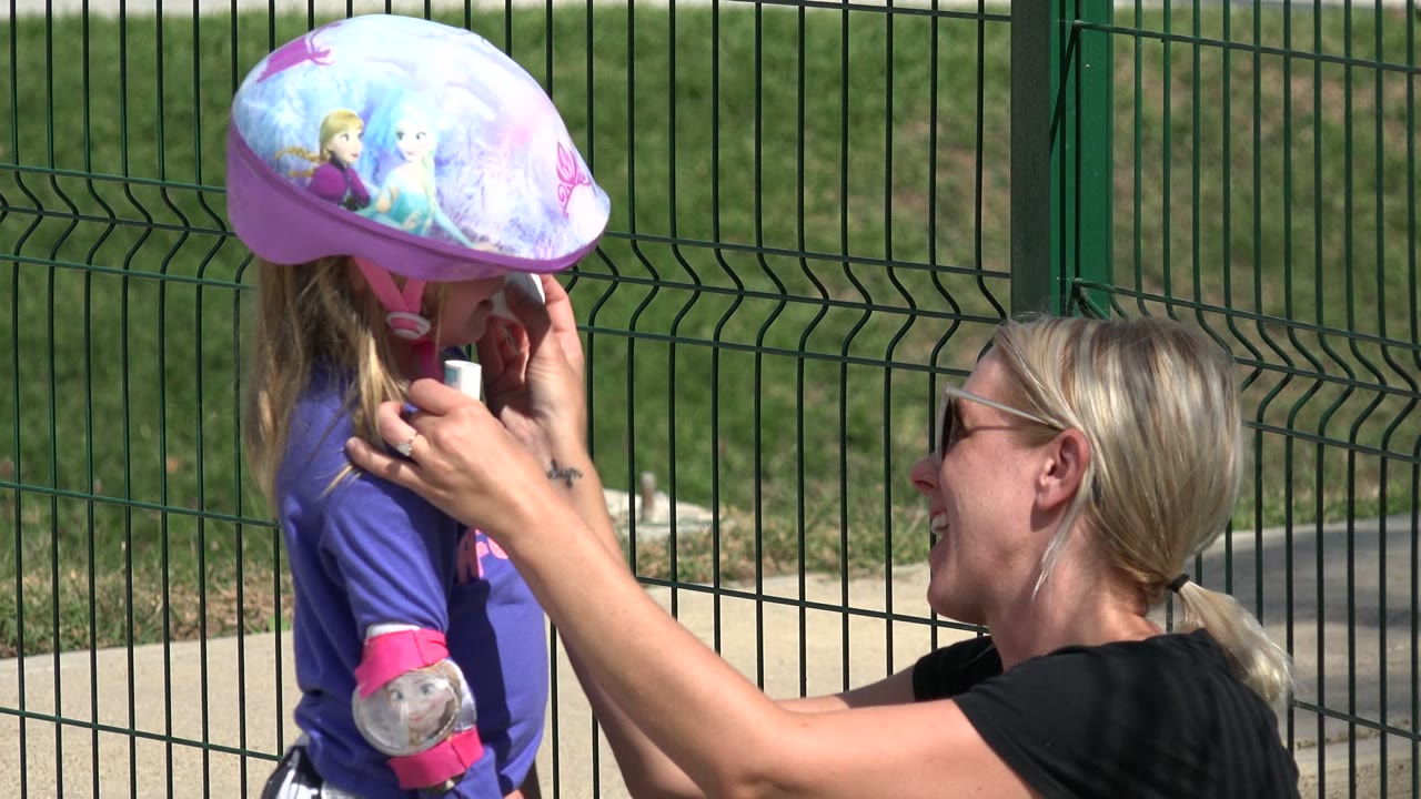 Woman adjusts protection on a helmeted child beside a green fence.