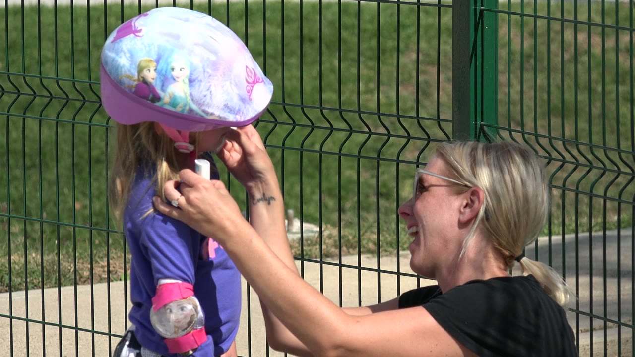 At 0:01, the mother smiles toward the child while both hands are near the girl's helmet strap and face, with bright sun casting sharp outdoor shadows.