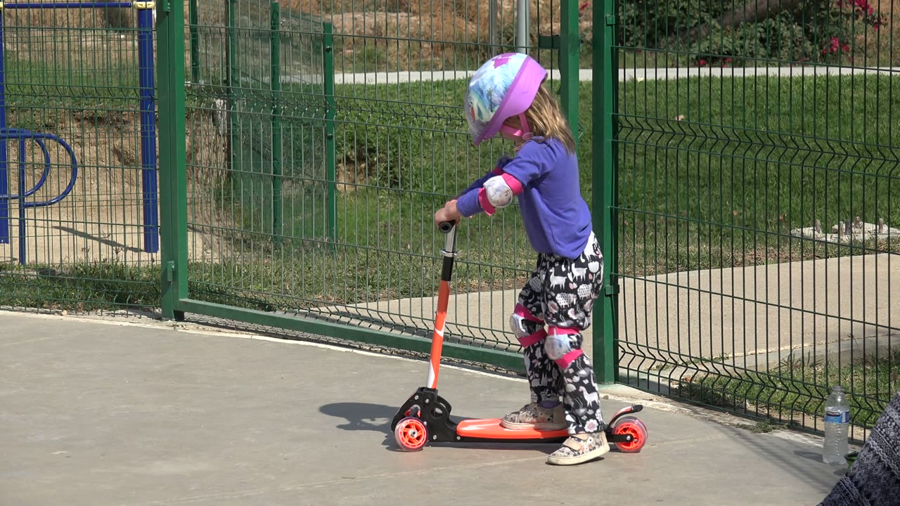 Little girl in a purple helmet standing on an orange scooter near a green fence.