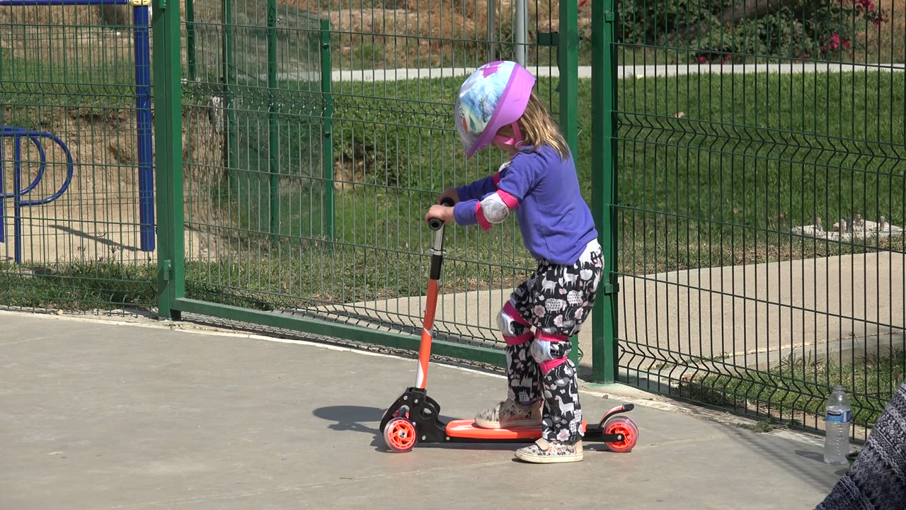 The child leans over the handlebar of the orange scooter in Los Cabos, Mexico, with bright sun on the concrete and a green fence in the background.