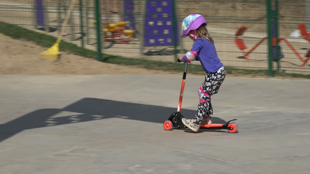 Young girl in helmet riding an orange scooter on a concrete playground path.