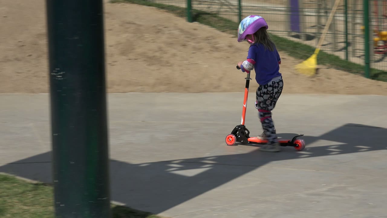 One second in, the girl continues riding the orange scooter along the concrete in Los Cabos, Mexico, with a grass edge, sand, and fencing behind her.