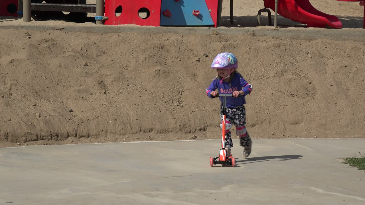 Young girl in a helmet riding an orange scooter on pavement.