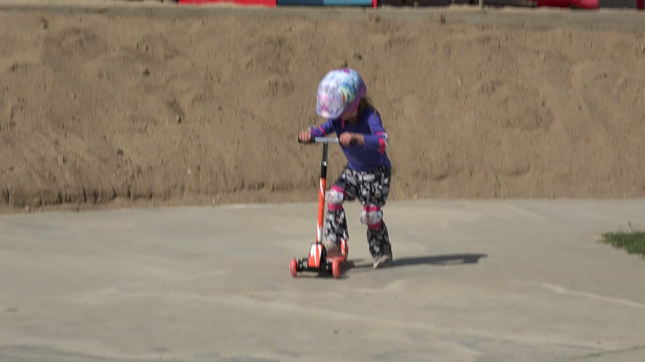 The girl leans over the scooter handlebars on a concrete path in Los Cabos, Mexico, with the sandy slope filling most of the background.