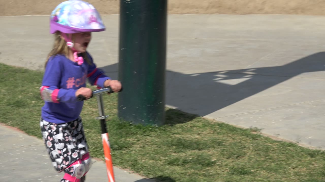 A young girl wearing a pink helmet and protective pads rides an orange scooter along grass and pavement in Los Cabos, Mexico.
