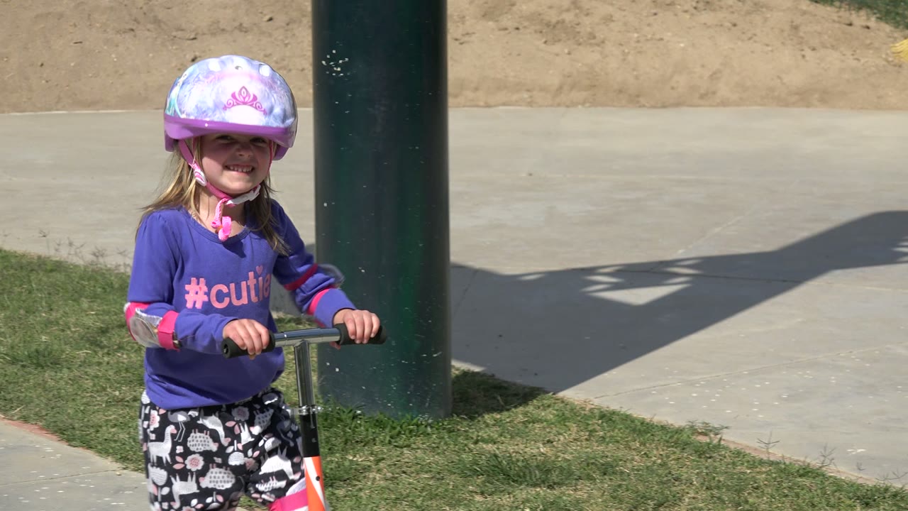 The child smiles while holding the scooter handlebars beside a dark green post, with grass and a concrete walkway behind her in Los Cabos, Mexico.