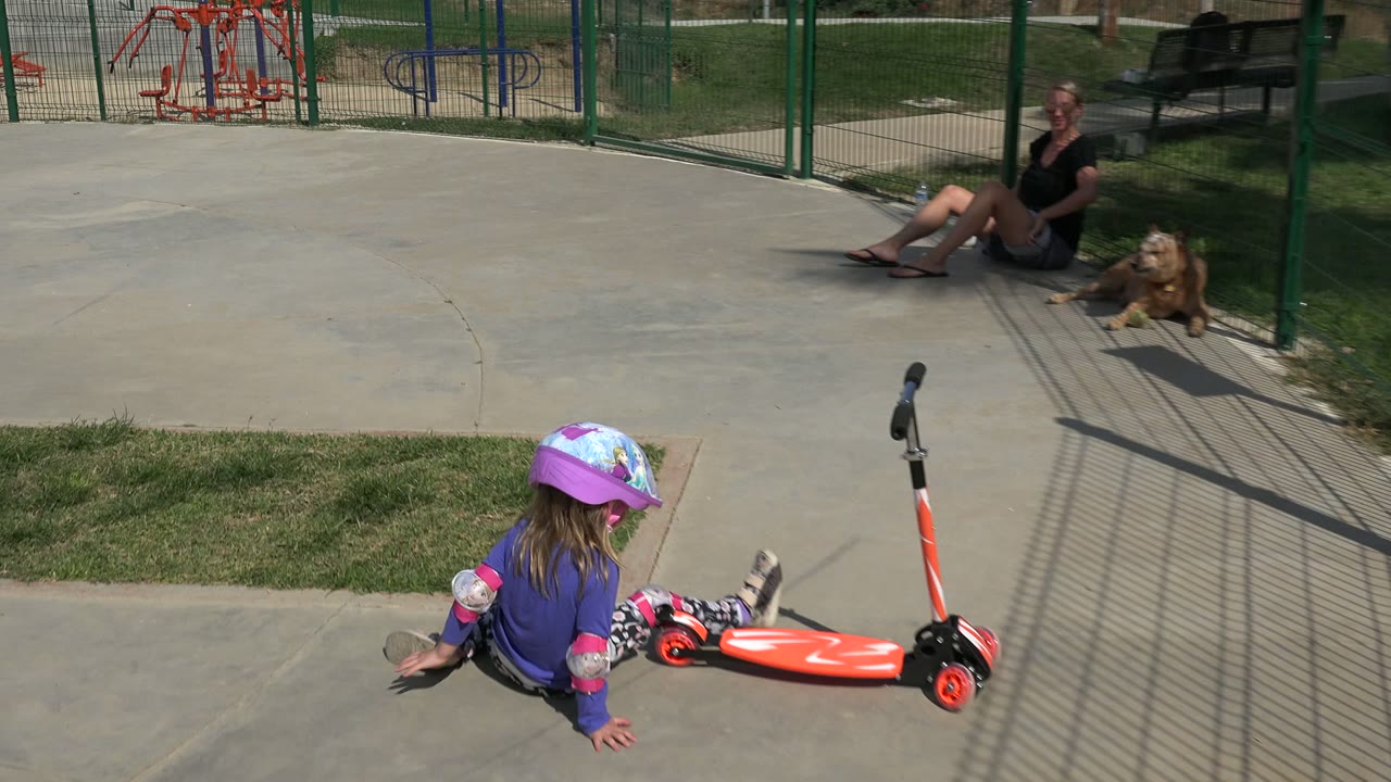 A young girl in a purple helmet sits on the concrete beside an orange scooter, with a woman and brown dog resting near a green fence in Los Cabos, Mexico.