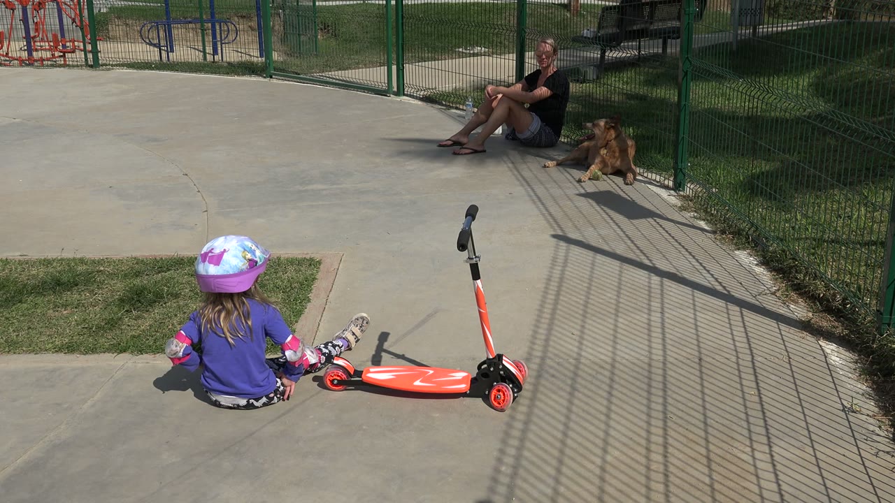 A young girl in a pink helmet sits with her back partly to camera beside a red toy scooter in a Los Cabos park, with a green fence, grass, an adult, and a dog behind her.