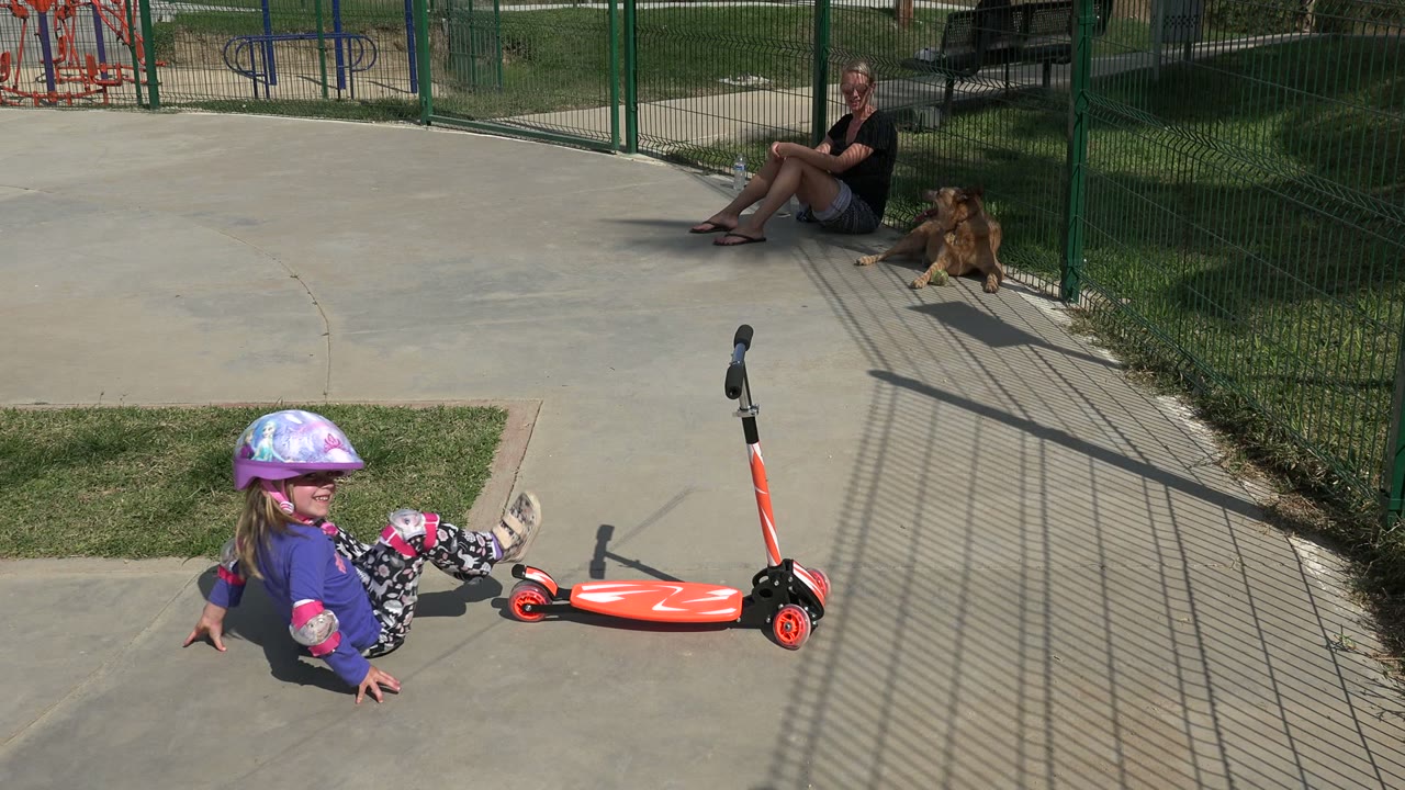 The helmeted girl sits on the concrete in Los Cabos and turns toward the camera, with the red scooter on its side and a fenced park area in the background.