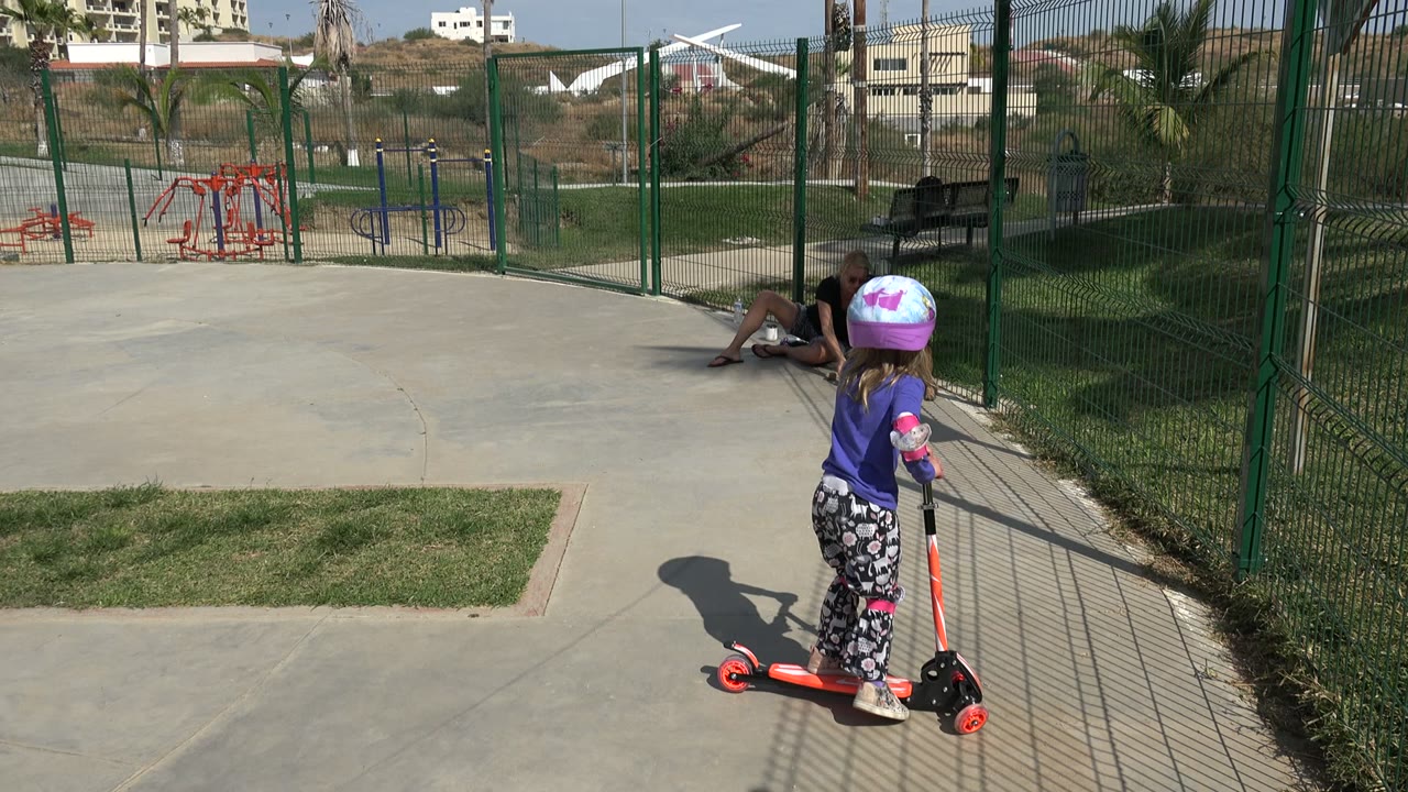 Young girl on an orange kick scooter near a green fence