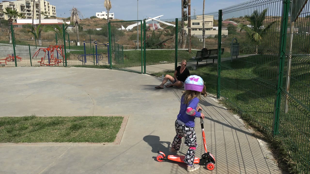 The child rolls forward on the orange kick scooter along the paved path in Los Cabos, passing fence shadows with playground equipment and buildings beyond the fence.