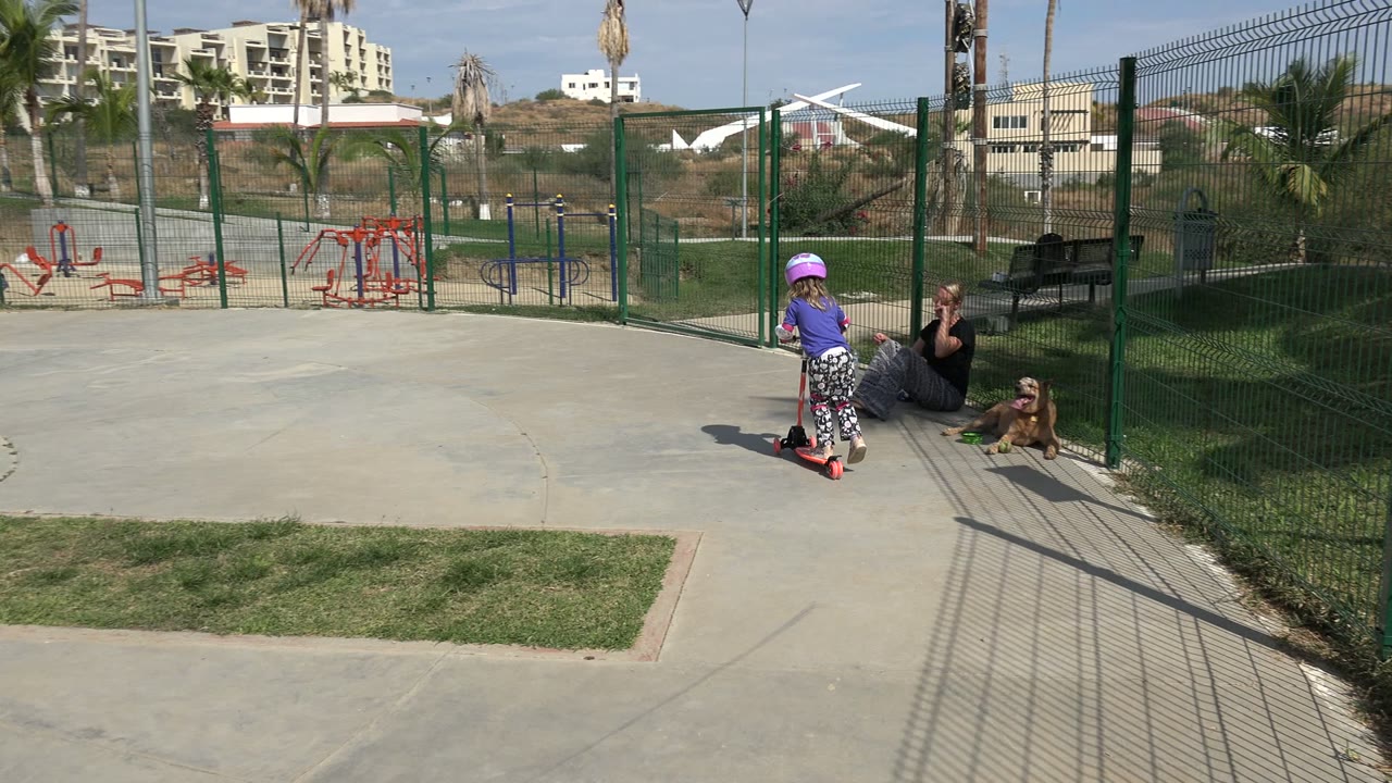 At the start of the Los Cabos park clip, a helmeted child rides a scooter on a concrete path while an adult sits beside a brown dog near a green fence.