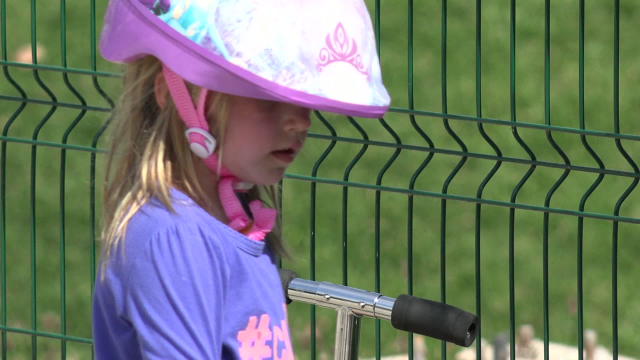 The child in a pink helmet appears more centered beside the chrome scooter handlebar, with the green fence and grass behind them in Los Cabos, Mexico.