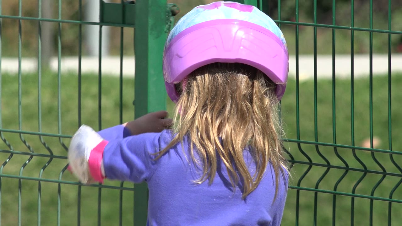 Young girl in a pink helmet facing a green metal gate.