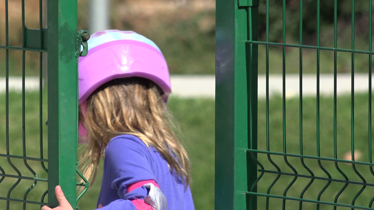 The child in a pink helmet moves beside a green gate post, with the vertical fence bars cutting across the Los Cabos park background.