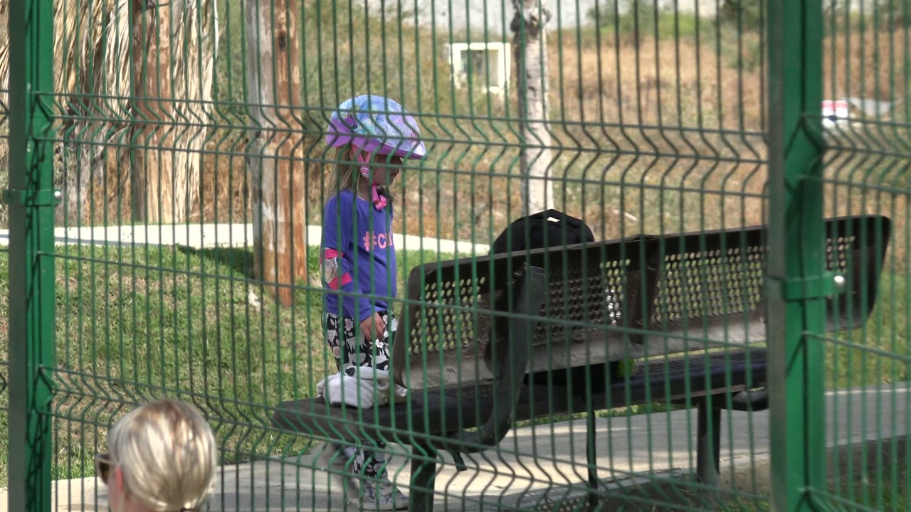 Young girl in a helmet behind a green metal fence near a bench.