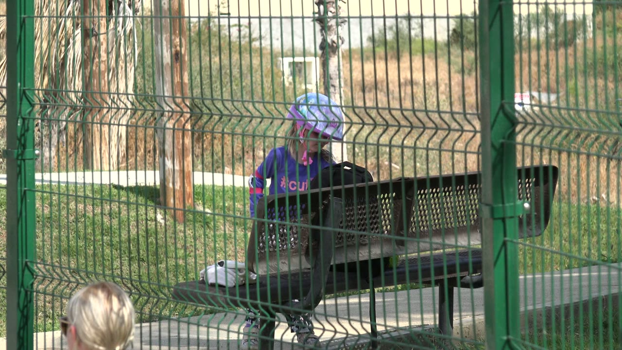 Helmeted girl stands closer to the gate area in Los Cabos, Mexico, with a white object on the ground near the black bench behind the fence.