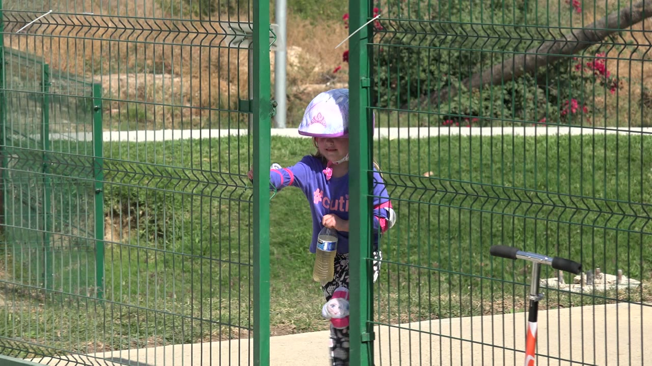 Child partly hidden behind a green park gate with a scooter nearby.