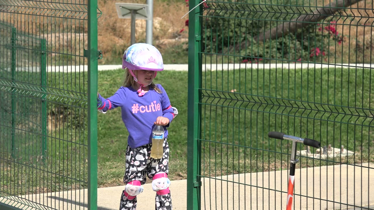 In Los Cabos, Mexico, the child opens the green park gate, wearing protective pads and holding a drink bottle beside the scooter.